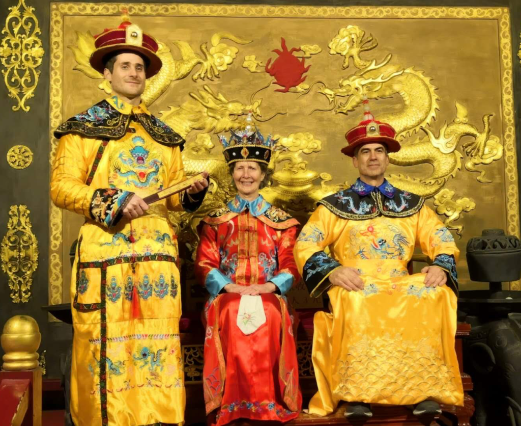 Chuck, his wife, and his son are dressed in traditional Chinese royal gowns staged in a throne room displaying a bright gold backdrop artistically embossed with dragons. Chuck's wife is seated between them, wearing a crown and dressed in a bright red gown as they roleplay a royal empress and her two subjects.
