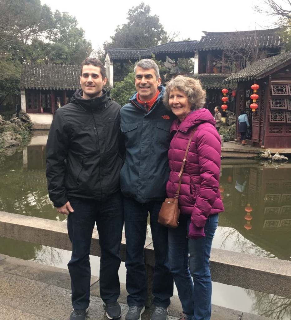 Chuck is posed between his wife and son on a stone trail in front of a Chinese garden pond surrounding a beautiful rufescent-stained wood building that features traditional Chinese architecture.