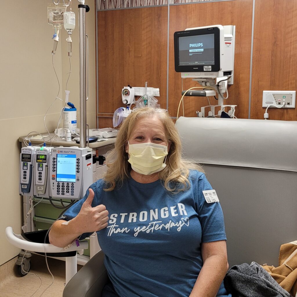 Dana is pictured in a patient chair while receiving an infusion. She is wearing a blue shirt, a mask, and appears to be in high spirits by the happy expression of her eyes and by the thumbs up she gives to the camera.