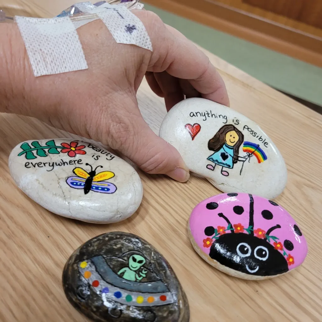A close up photo of four of Dana's painted rocks. Dana has an IV in her left hand while she picks up one of the rocks to show to the camera. The first rock reads "everything is beautiful" in black paint with a blue and yellow winged butterfly and a red flower painted next to the words. The next rock shows an green alien throwing up a peace sign while manning a spaceship with rainbow lights. The rock above that is an incredibly cute rock painted as a pink ladybug wearing a red flower necklace. The fourth painted rock that Dana is displaying in her hand shows a girl with long brown hair holding string tied around a small rainbow that is floating next to her like a balloon. This last rock reads "anything is possible" in black paint above a red heart.