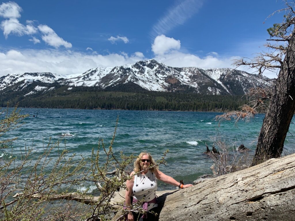 Dana is standing against a gigantic log of driftwood in the forefront of a beautiful landscape. Behind her, gentle waves of the blue-green river are floating by, while sprawling snowy mountains stand in the background.