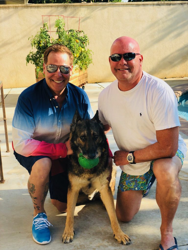 Jim and Kevin nealing down with their dog smiling.
