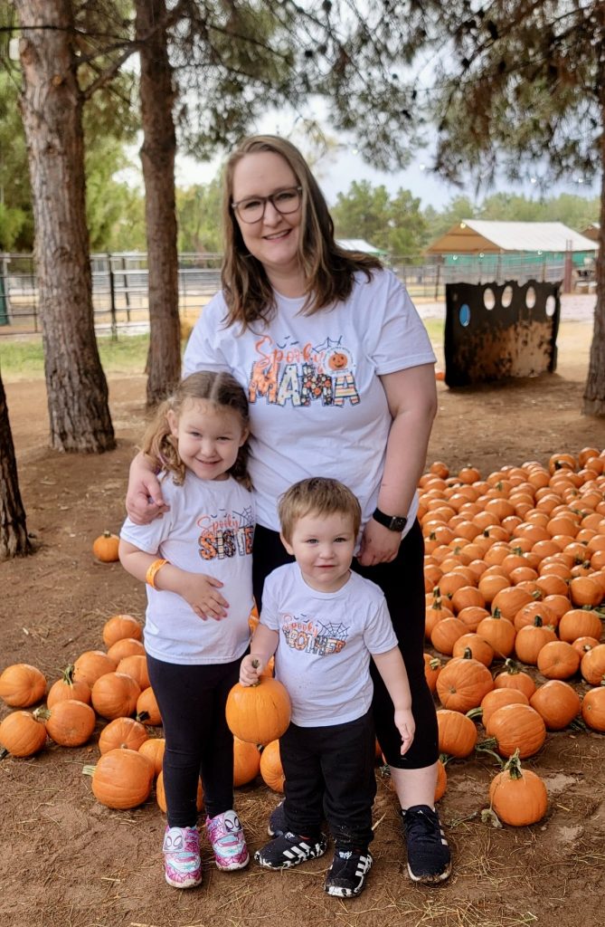 Alexandra is standing in front of dozens of small pumpkins on the ground outside while posing with her son and daughter. It appears to be halloween season in the Fall and in the background is a farm with gates, tall pine trees and hay-covered soil. She has medium-length blonde hair, large brown-framed glasses and is wearing black leggings and black athletic shoes. All three of them are wearing matching white halloween-themed T-shirts. Alexandra's says "Spooky Mama", and her son and daughter's shirts say "Spooky Brother" and "spooky sister".