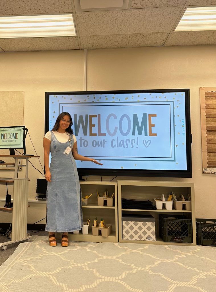 Esmeralda, standing at the front of a warm and inviting classroom, gestures proudly to the image on the projector display that reads "Welcome to our class!". Esmeralda is smiling, wearing large-framed, stylish and transparent glasses, with smooth styled warm-brown hair that frames her face with soft waves. She is wearing a modest white t-shirt under a long, muted light-blue denim dress with tall clog-heels with wide brown straps.