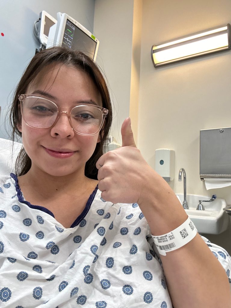 Esmeralda is laying in a hospital bed wearing a hospital gown and her stylish large-framed transparent glasses. She is cheerfully and encouragingly holding a thumbs-up while softly smiling at the camera as she takes a self-portrait.