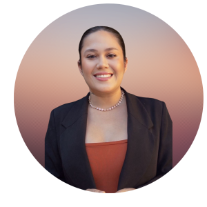 Angela has dark hair pulled back, wearing a black blazer over a rust-colored top and a beaded necklace, smiles at the camera. The circular frame of warm tones reflects the welcoming spirit of About The MOG Project.
