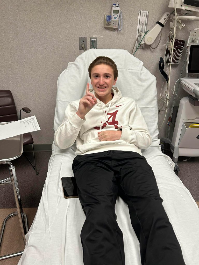 A young man in a white hoodie with a red A logo lies on a hospital bed, smiling and holding up one finger after receiving a MOGAD diagnosis. Medical equipment is visible in the background.