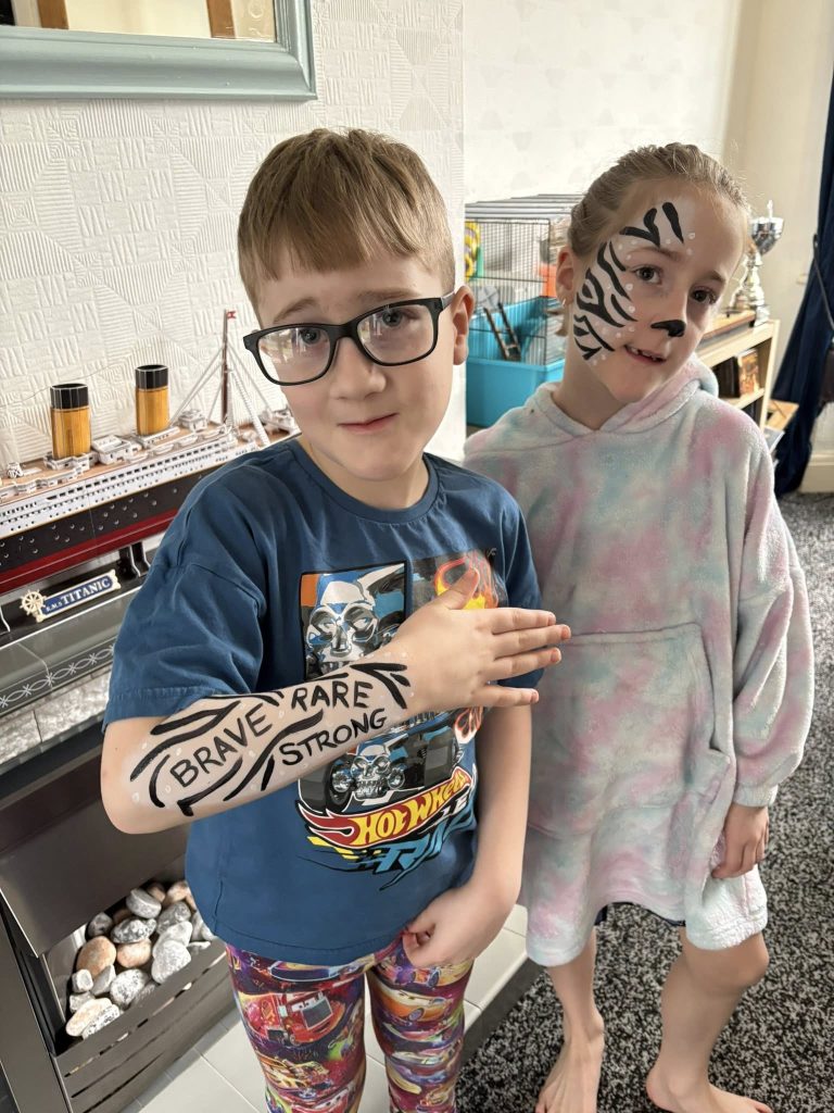 Two children stand indoors. One boy, recently given a neurological diagnosis, wears glasses, a blue T-shirt, and patterned shorts with BRAVE, RARE, STRONG written on his arm. A girl in a hoodie stands beside him; both have faces painted with tiger stripes.