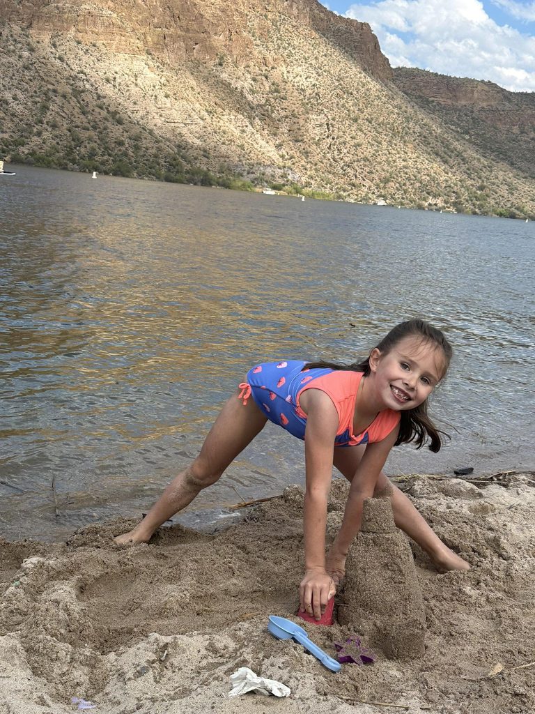 A young girl in a blue and pink swimsuit smiles while playing in the sand by a lake, with mountains and a partly cloudy sky in the background. She is leaning forward, using a red mold to shape the sand, her spirit bright despite her recent MOGAD diagnosis.