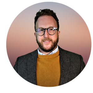 A man with short brown hair, glasses, and a beard is wearing a brown blazer over a mustard sweater and collared shirt, standing in front of a circular gradient background—perfect for an About The MOG Project profile.