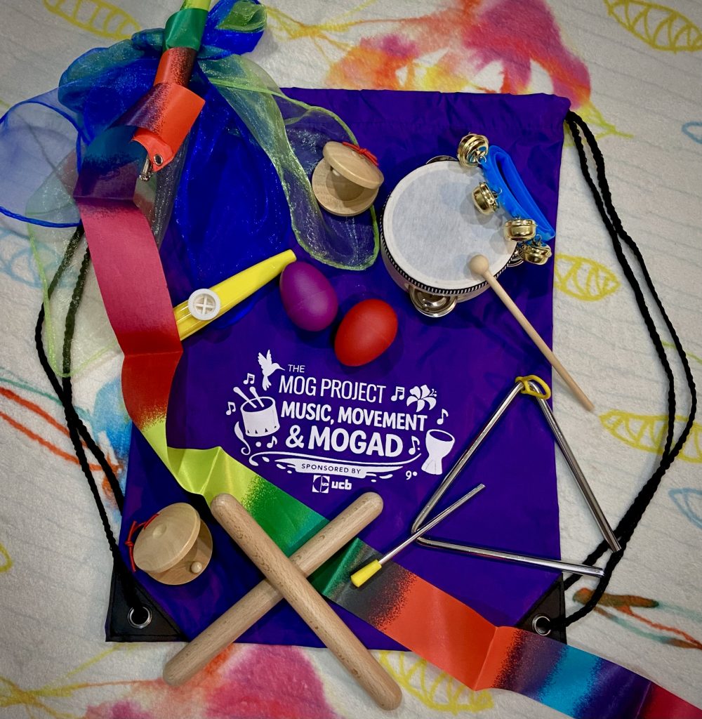 A purple drawstring bag labeled The MOG Project: Music, Movement, & MOGAD is surrounded by colorful scarves, wooden rhythm sticks, egg shakers, a tambourine, triangle, and a wooden castanet—perfect for children to explore music on a bright patterned background.