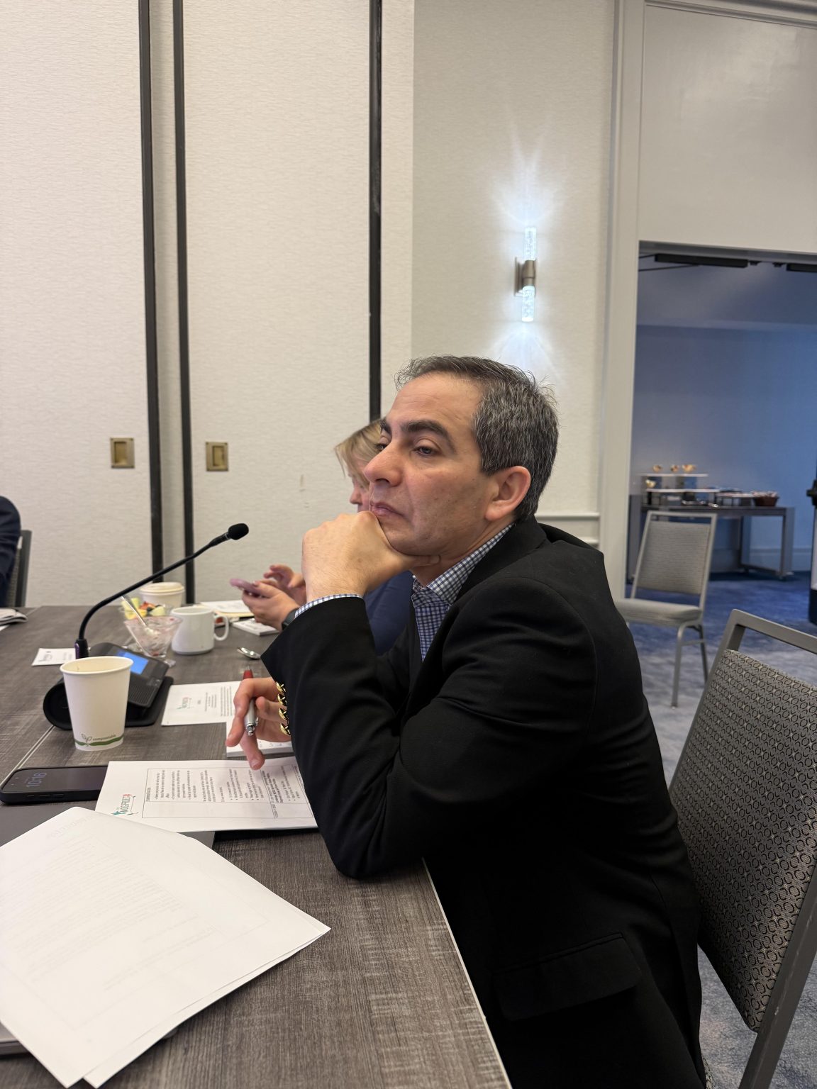 A man in a suit sits at a conference table with papers, a microphone, and a coffee cup in front of him. He is resting his chin on his hand and appears to be listening attentively.