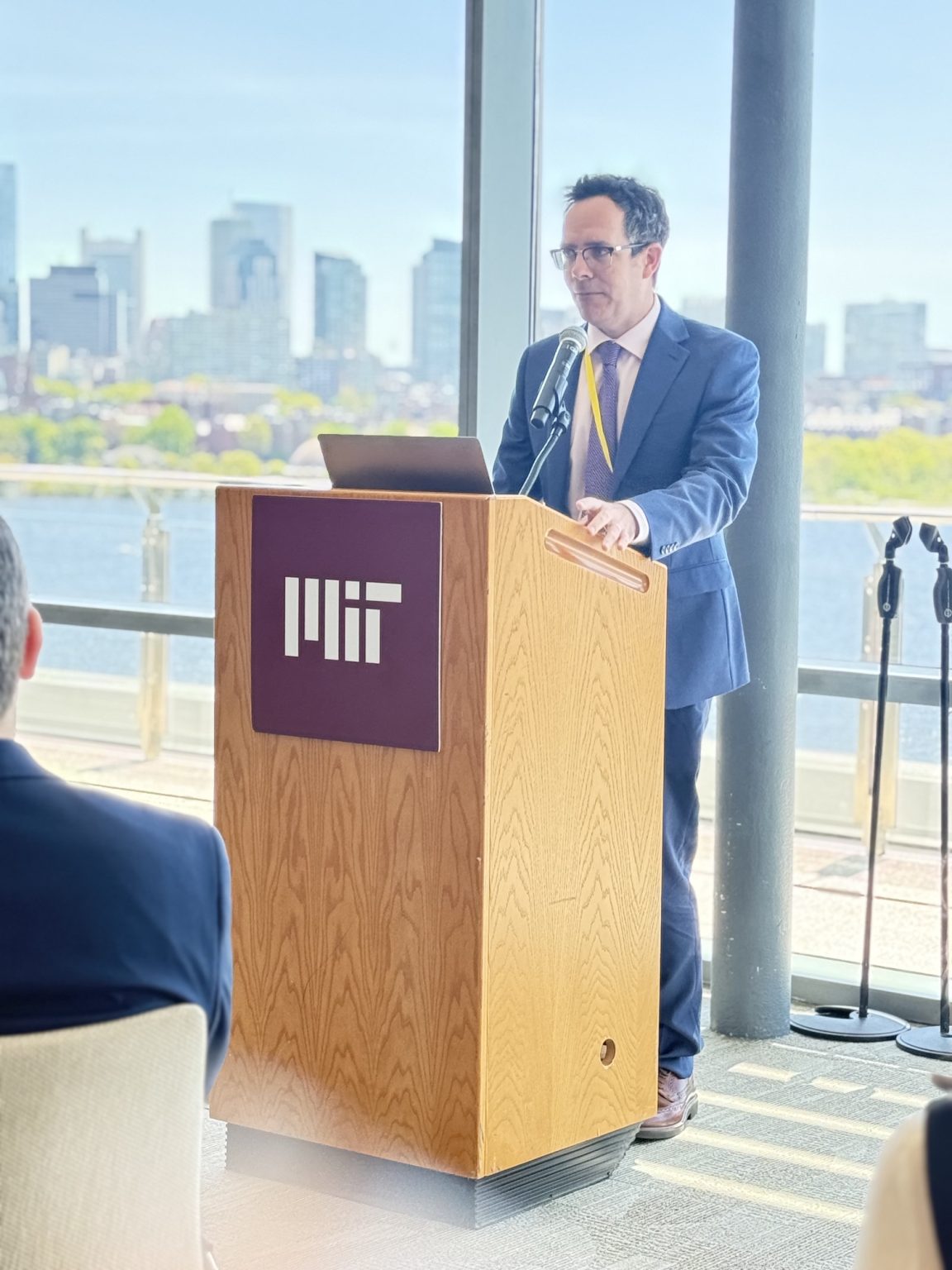 A man in a suit speaks at a podium with the MIT logo, addressing an audience in a bright room with large windows overlooking a city skyline.