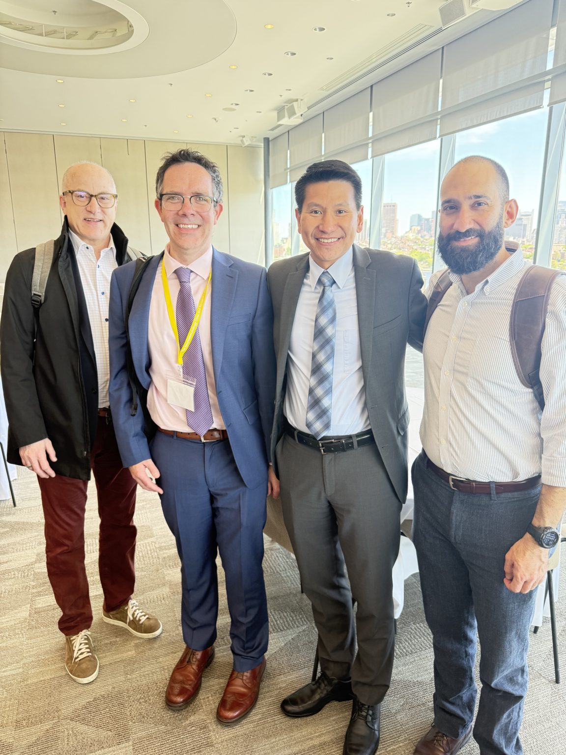 Four men stand together indoors, smiling at the camera. Two are wearing suits with ties and name badges, and the other two are dressed in casual shirts and slacks with backpacks. Large windows show a city view in the background.