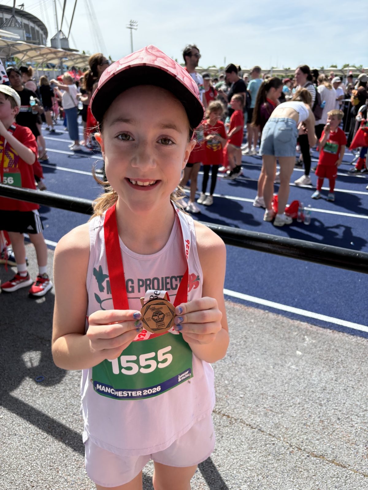 A smiling young girl wearing a red cap and race bib 1555 proudly holds up a finisher’s medal. She stands on a track surrounded by other children at the busy MOGAD fundraiser event in Manchester 2024.