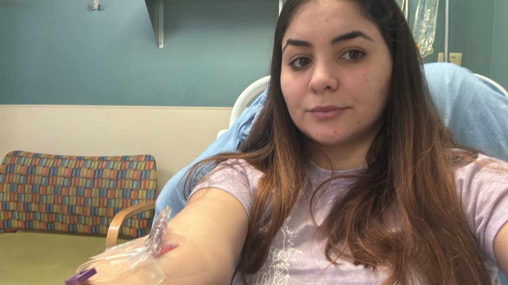 A woman with long brown hair sits in a medical setting, wearing a light purple shirt, with an IV inserted in her arm. She looks at the camera with a neutral expression.