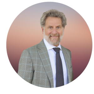 Scott with curly gray hair and a beard smiles, wearing a checked gray suit, white shirt, and dark patterned tie, posed in front of a circular background with a gradient from pink to purple.