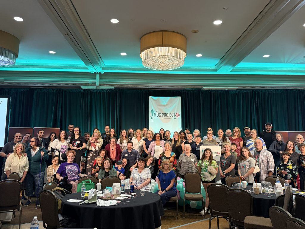 Group photo of people in a conference room under a MOG Project sign at the MOGAD Patient Summit 2026, with tables, chairs, and green lighting in the background.